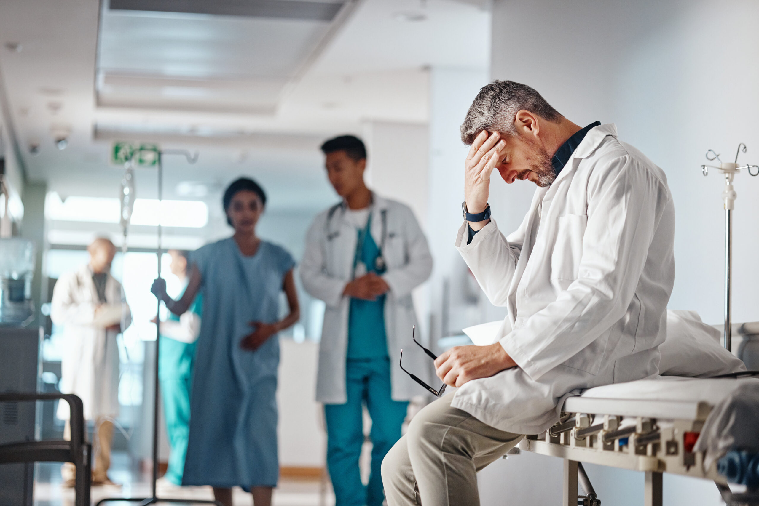 Doctor in a white coat sits on a hospital bed, hand to forehead, looking stressed while colleagues stand in the background.
