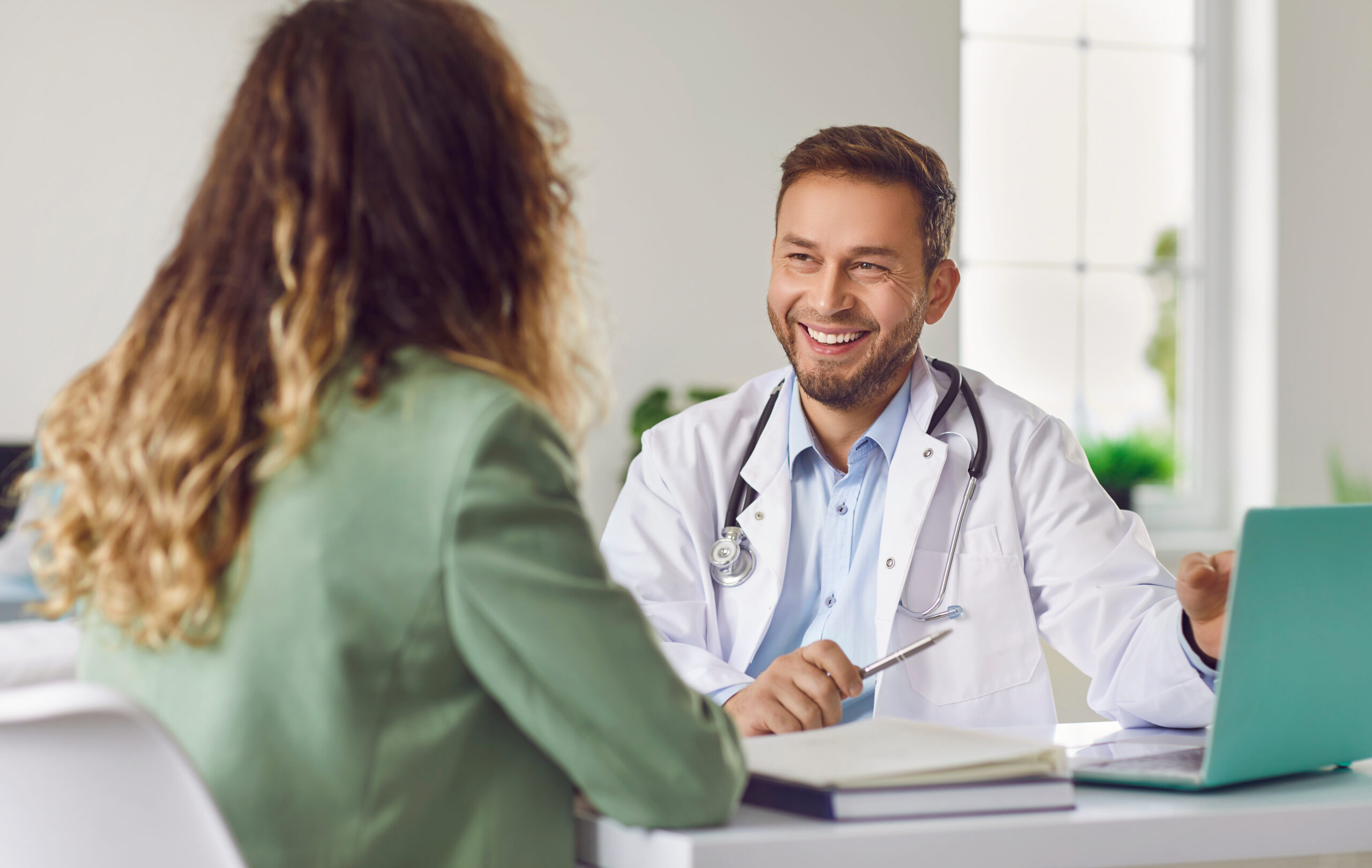 Smiling doctor in a white coat consults with a patient across a desk in a clinic, stethoscope visible.