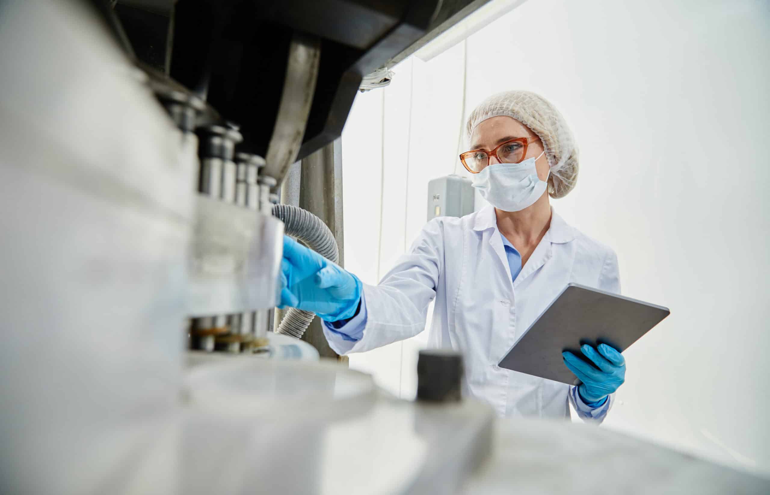 Scientist in a lab coat, mask, hairnet, and blue gloves inspecting machinery with a clipboard nearby in a clean lab setting.