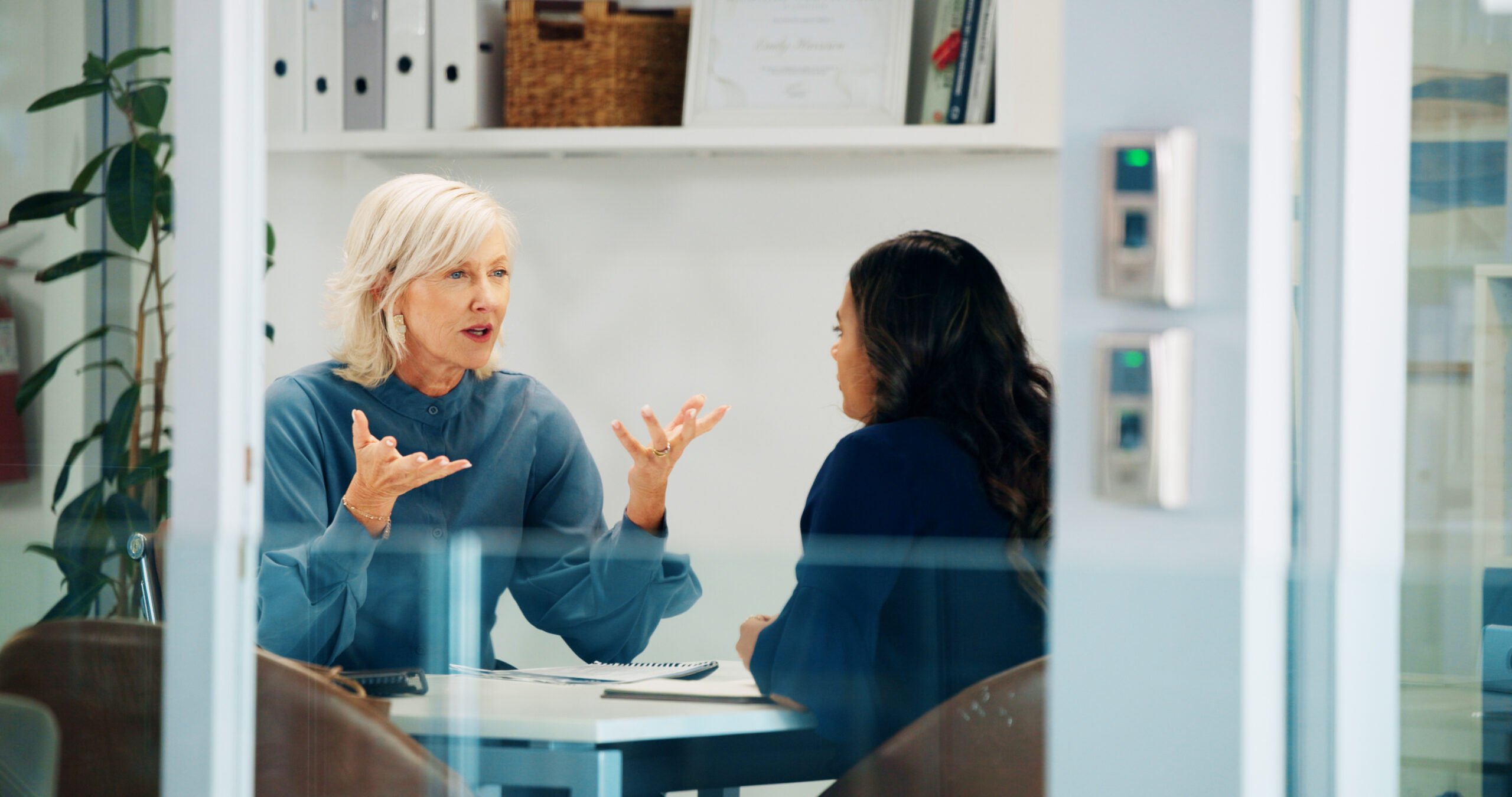 Two women sit at a glass-walled office desk having a serious conversation, one with short white hair gesturing with her hands.
