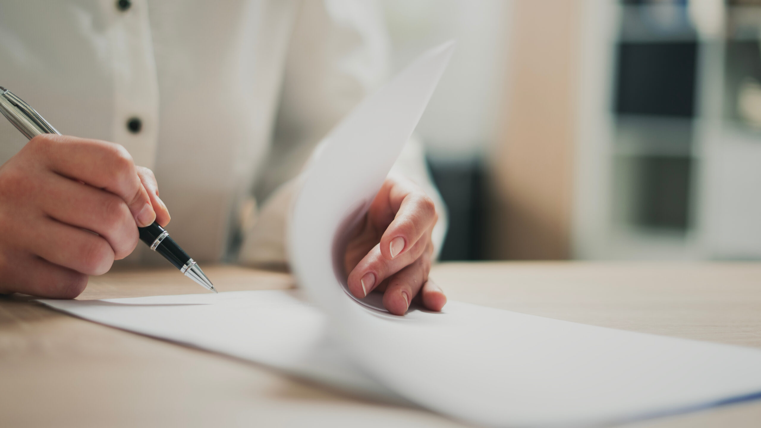 Close-up of hands signing a document with a silver pen on a wooden desk, papers nearby.