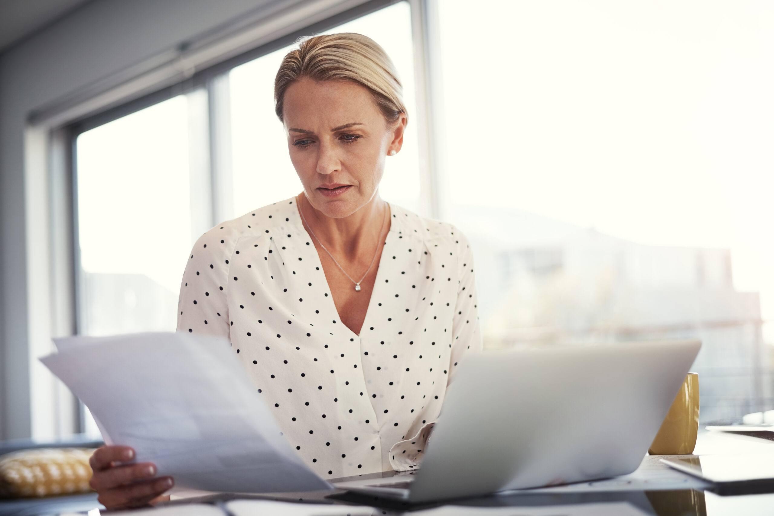 Woman in a white polka-dot blouse reviews papers at a desk by a window, with a laptop nearby.