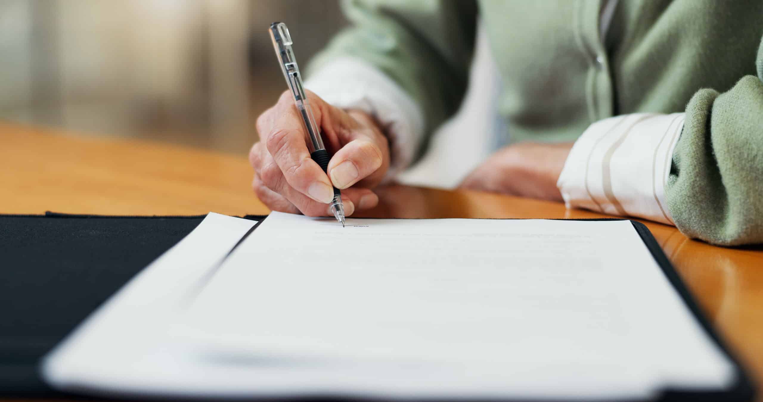 Close-up of a hand signing a document with a pen on a wooden desk, focus on the pen tip touching paper.