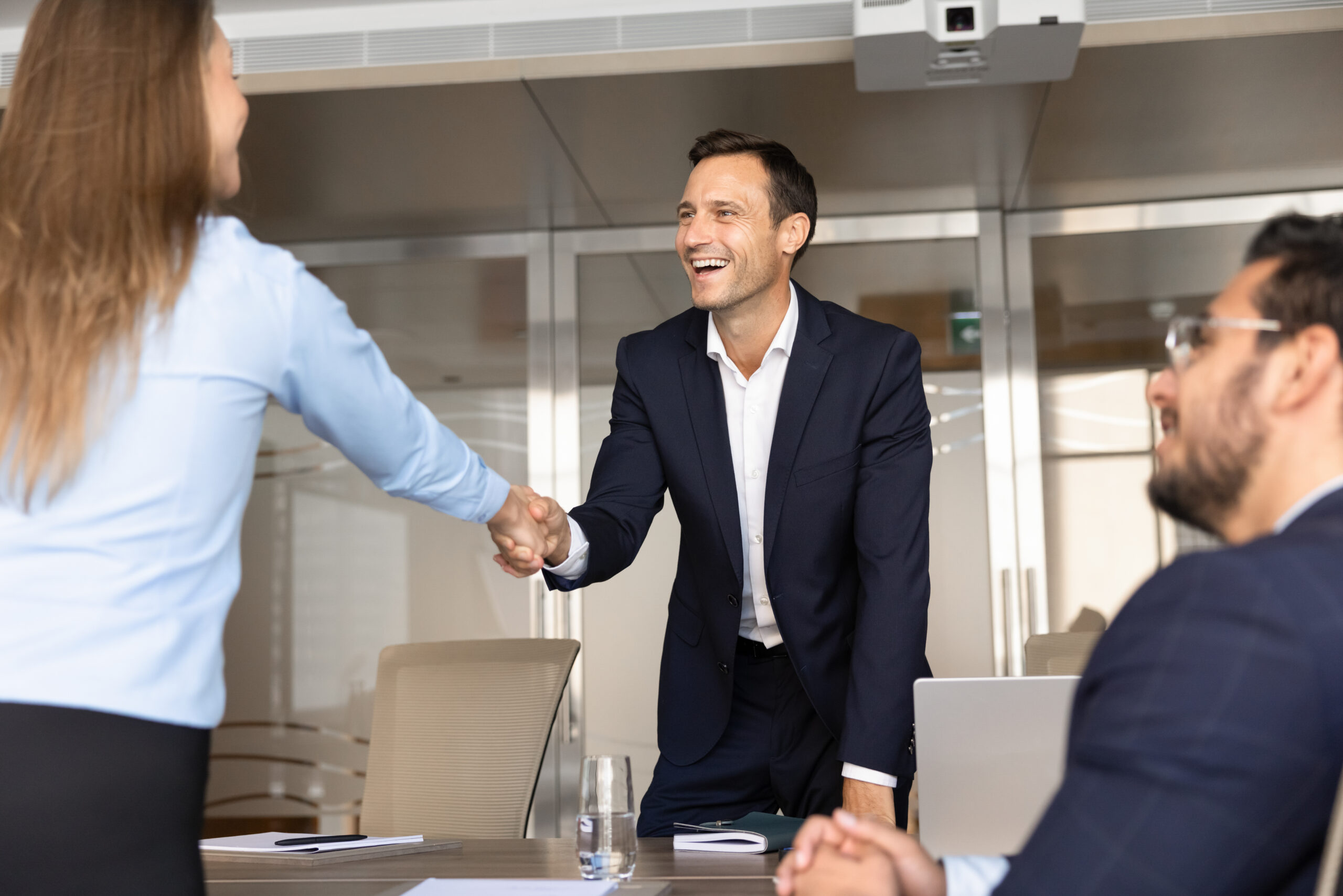 Two coworkers shake hands across a conference table while smiling in a modern office setting.