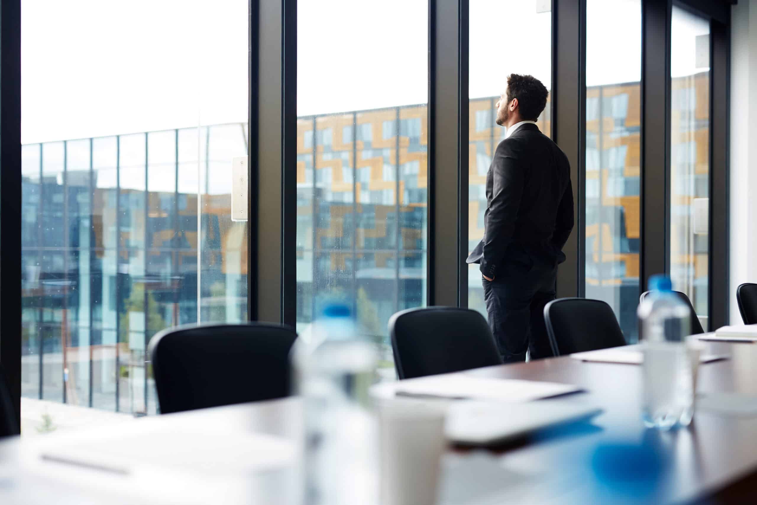 Man in a dark suit stands by large floor-to-ceiling windows in a modern conference room, looking outside.