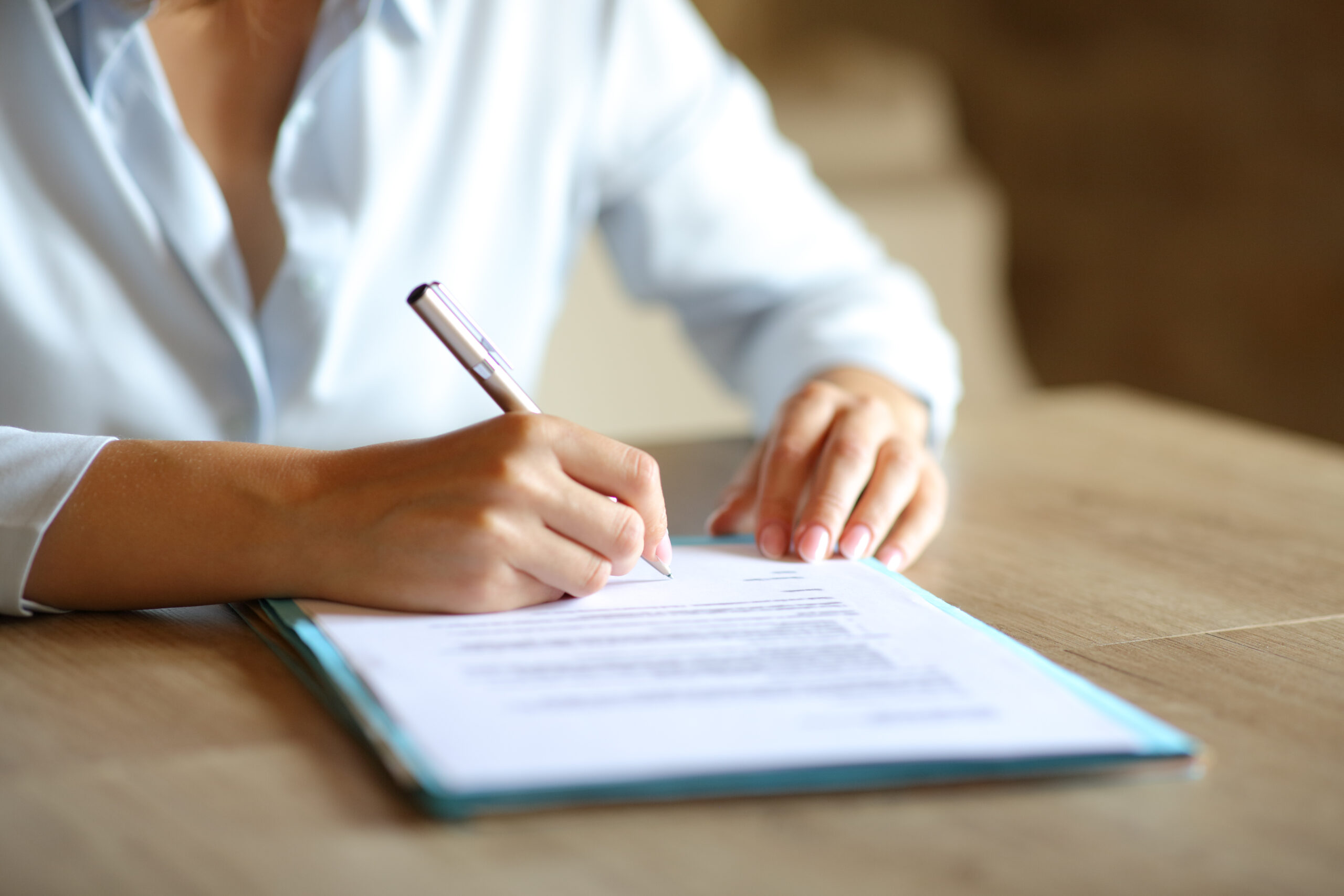 Person's hands writing with a pen on a document spread out on a wooden desk.